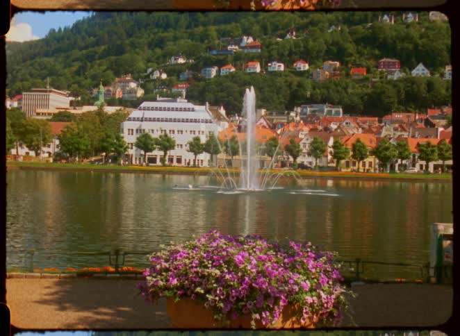 A fountain sprays in a lake surrounded by buildings and hills