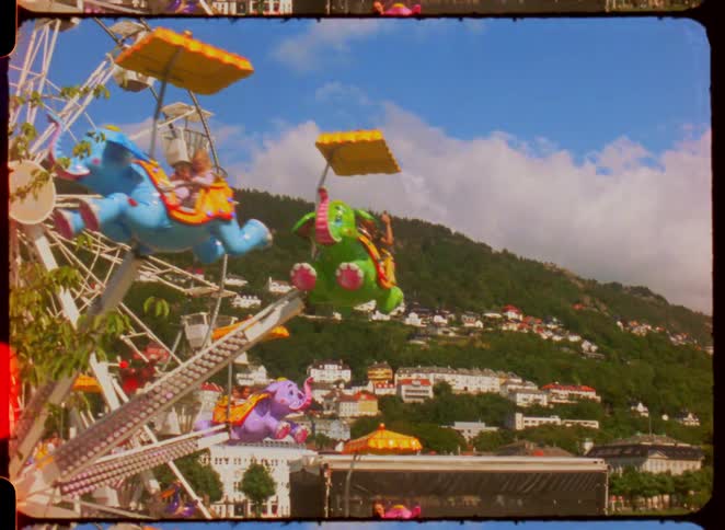 A vibrant amusement park scene unfolds with a ferris wheel featuring elephant shaped rides carrying people set against a backdrop of a hilly town under a bright blue sky