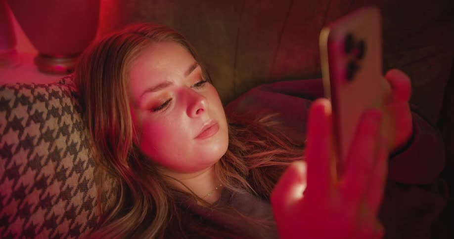The video shows a young woman sitting on a couch and holding her phone the room is dimly lit with a red glow suggesting it is nighttime