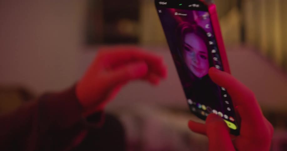 A hand holds a phone showing a live selfie its screen glowing magenta in a dim cozy room
