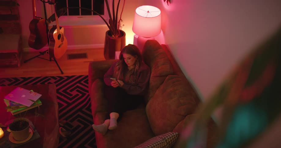 Woman sitting on a brown couch uses her phone in a room lit by a pink lamp with guitars resting against the wall