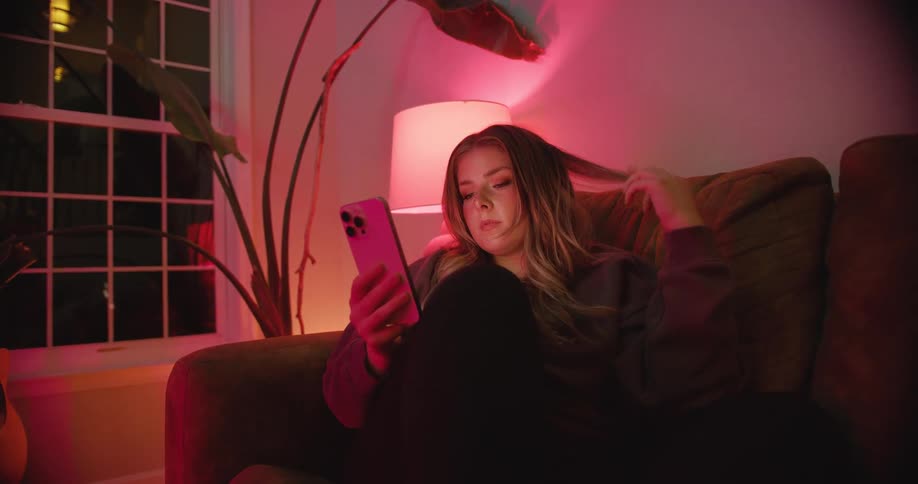 A young woman lounges on a sofa scrolling her phone under soft pink lamp light
