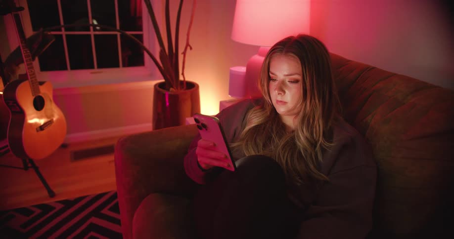 A young woman sitting on a couch in the living room at night using her mobile phone with a lit lamp beside her and a guitar on a rack near the window