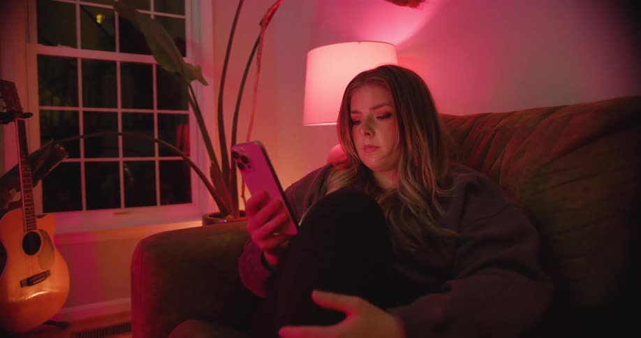 Young woman sitting on the sofa in a living room at night interacting with her smartphone