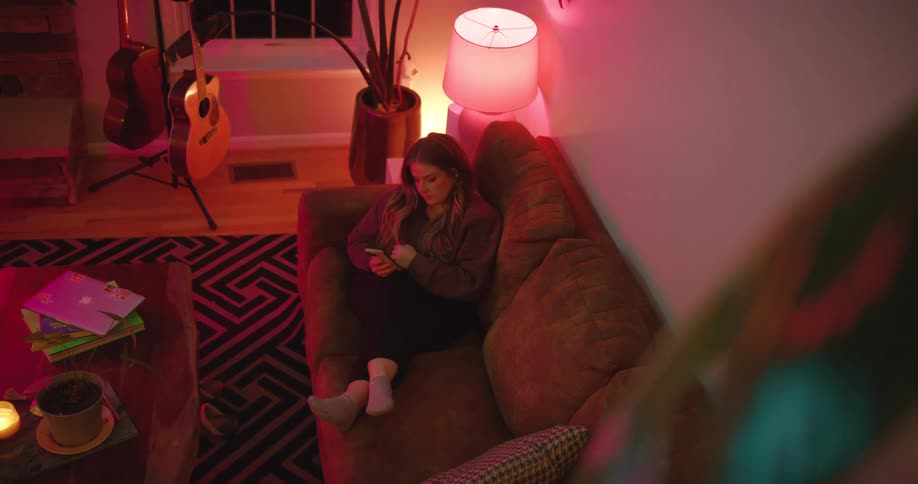 A young woman lying on a couch in the living room at night using her mobile phone with a lit lamp beside her and a guitar on a rack near the window
