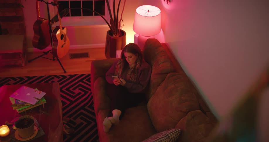 The video shows an overhead shot of a young woman lying on a brown sofa in a living room she is looking at her cell phone with both hands a guitar on a stand and a patterned rug are visible in the background