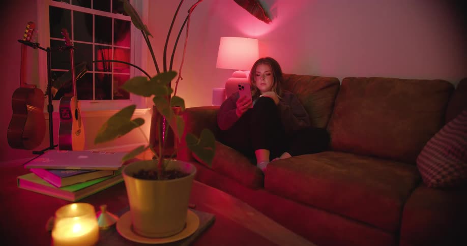 The video shows a young woman sitting on a brown couch in a dimly lit room with a pinkpurple ambiance she is engrossed in her phone which she holds with both hands in the foreground there is a potted plant on a coffee table next to a stack of books a