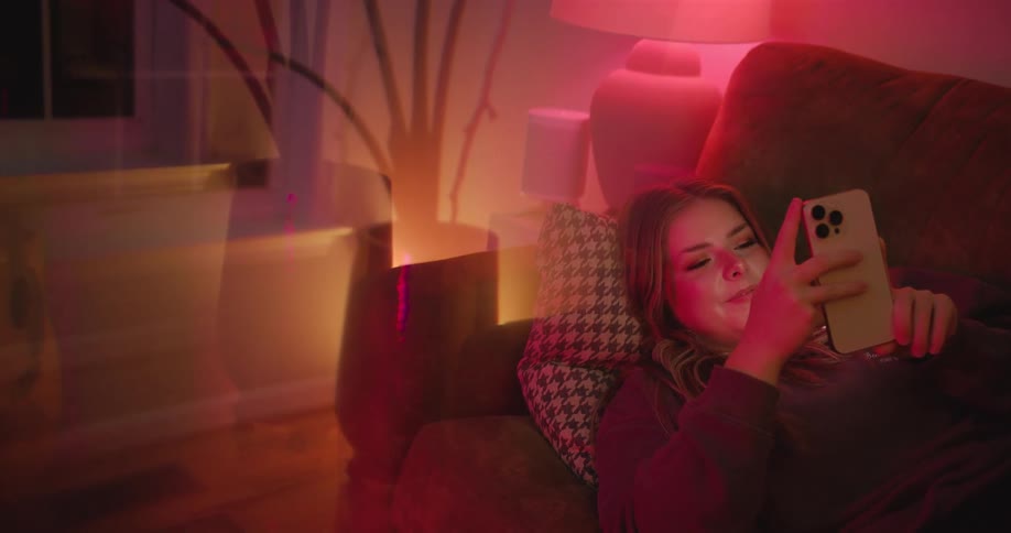 A young woman scrolls on her phone on a couch under red lighting