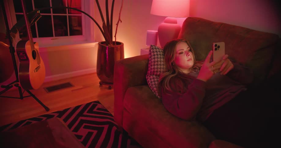 The video shows a young woman sitting on a couch in a dimly lit room scrolling through her cell phone she is illuminated by a warm pink light and at one point she smiles an acoustic guitar is visible on a stand behind her