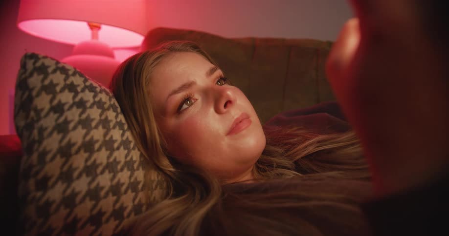 A woman reclines on a couch looking at a cell phone as a red lamp washes the room with evening warmth