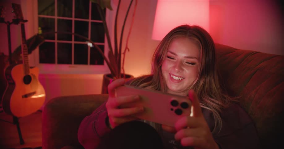 A woman with blonde hair sits on a couch in a dimly lit room holding a smartphone horizontally with both hands and smiling as she watches a video