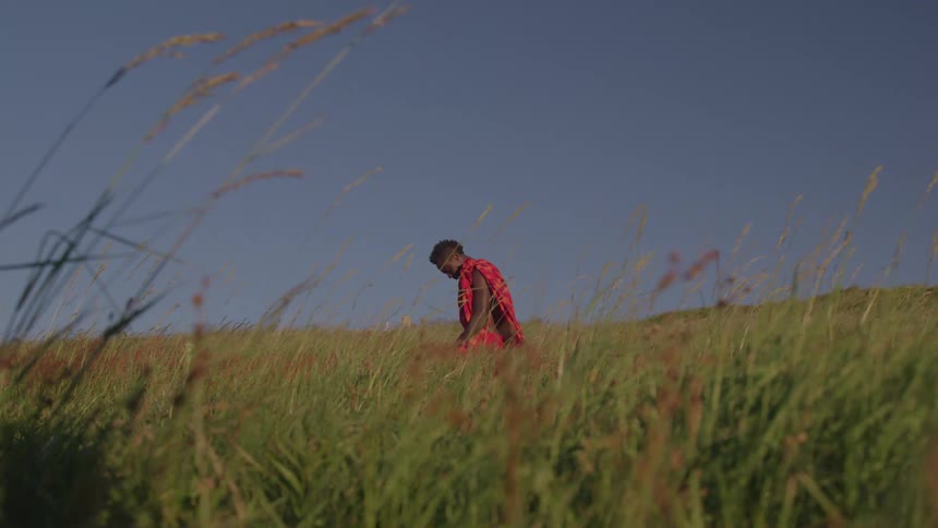 An african man wearing native attire kneels on a grassland on a sunny day
