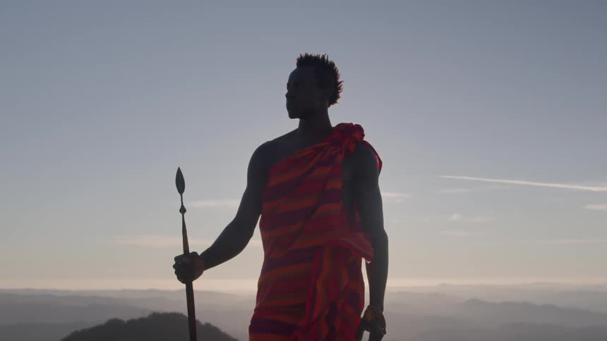 A man wearing a cloth suit and holding a spear is standing in an outdoor environment looking intently at the same position