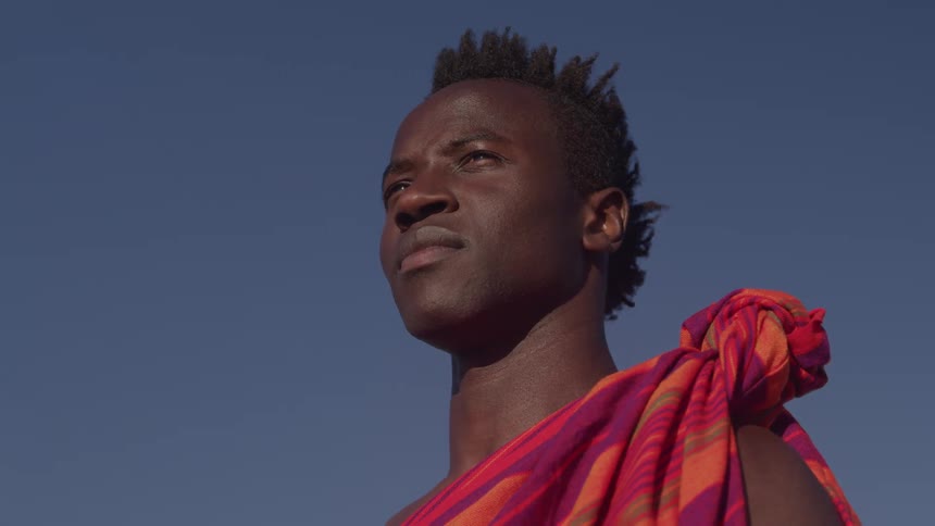 The face of a man wearing a red cloth tied around his shoulder and he is staring straight ahead in contrast to the blue sky behind