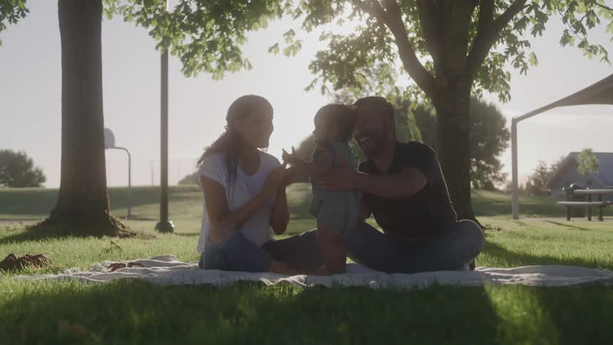 A family of three on a blanket over the grass in a park with the man and a woman playing with the baby that does their best to stay upright