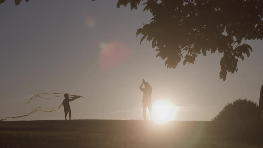 A man claps as a boy runs and tries to fly a kite as the sun goes down behind them