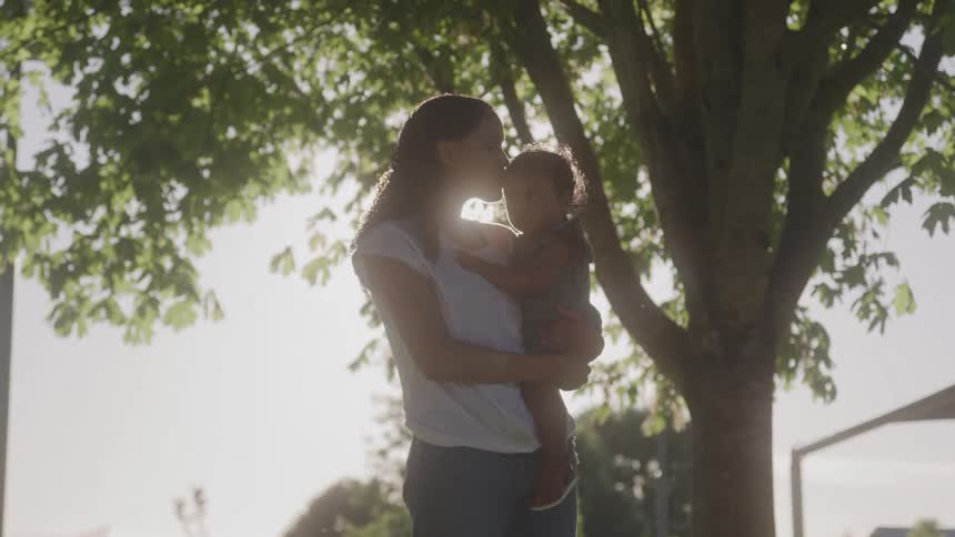 A woman holding her daughter in her arms and kissing her on the forehead in an outdoor setting next to a tree