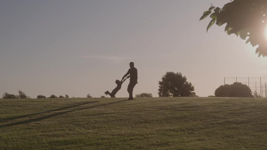 Man turning in place on a grassy slope in the park swings his son around on a sunny summer day