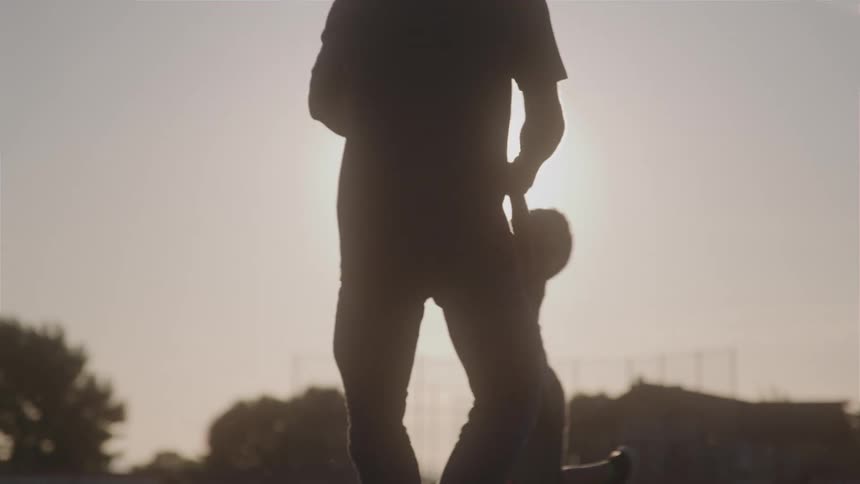 The silhouette of a father holding his son by the arms and spinning around and behind him there is the sun shining brightly