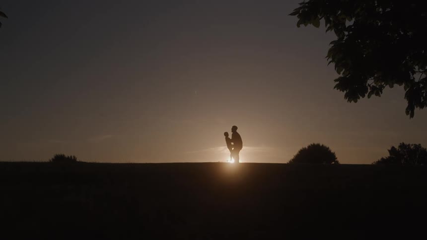The video shows a silhouette of a man and a child on a grassy hill against a sunset the man bends down to pick up the child then stands back up while holding them in his arms