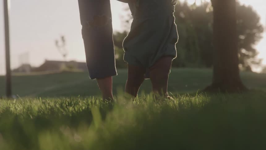 A babys feet as they try to take their first steps on a parks grass with a person in torn jeans helping from behind