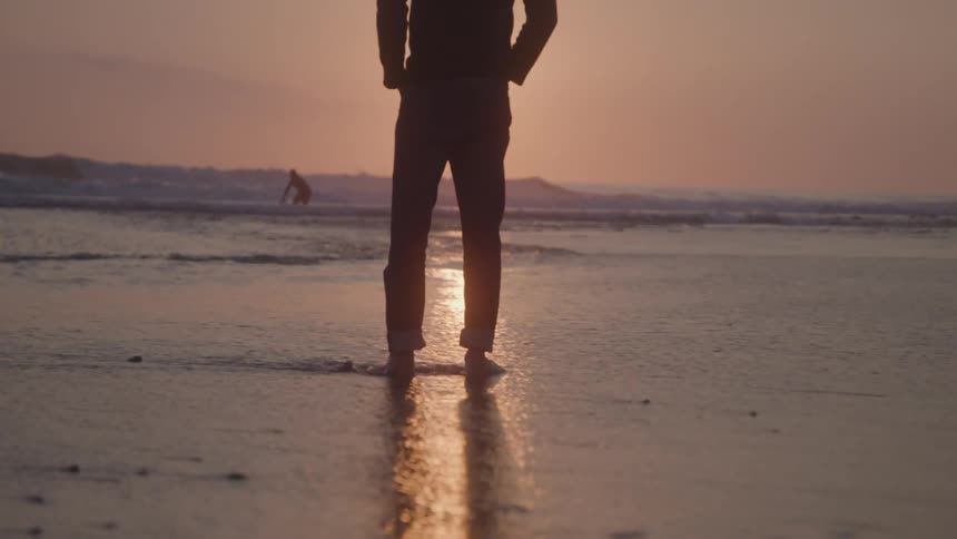 A man stands silhouetted on a beach at sunset with a person surfing in the background