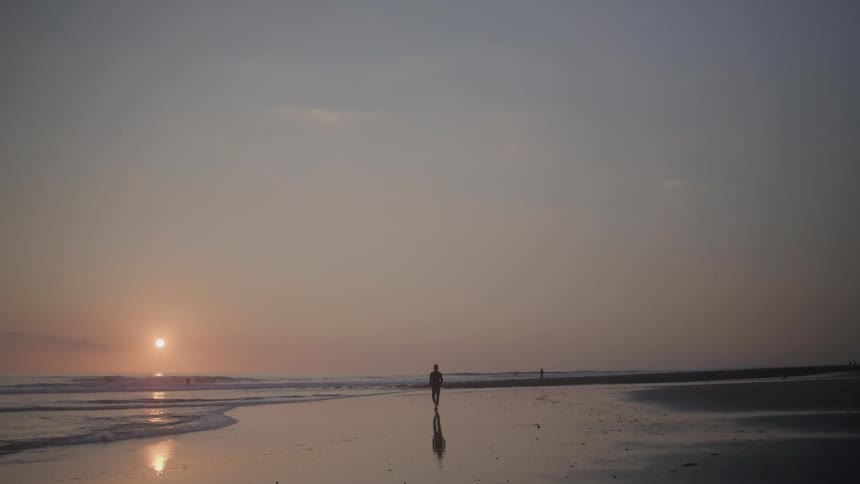 The video shows a wide shot of a beach at sunset a person stands in the distance silhouetted against the setting sun while small waves lap at the shore and create reflections on the wet sand