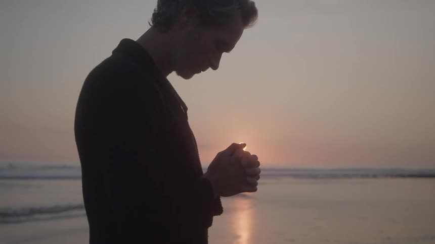 A man stands silhouetted against a sunset on a beach holding his hands together as if in prayer or contemplation the setting sun is a small bright orb of light just above his hands