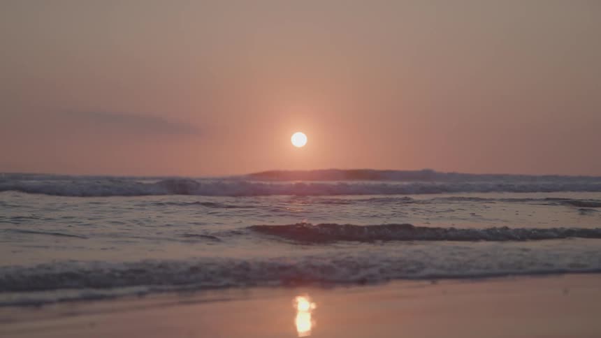 Surf rolling onto the beach covers the wet sand reflecting the sun sitting in the red sky