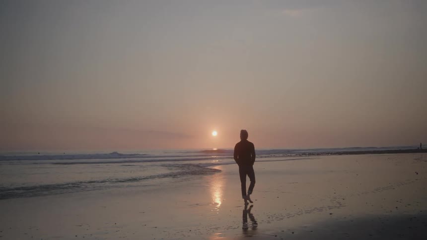 A man walks slowly along a sandy beach as the sun sets over the ocean his silhouette is reflected in the wet sand as waves roll gently onto the shore