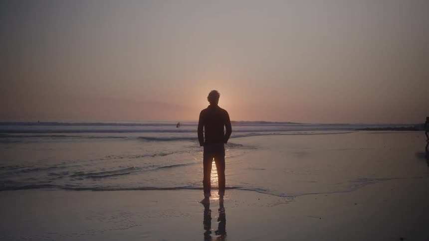 Man standing on the beach is silhouetted by the sun shining from the horizon while a surfer walks toward the water