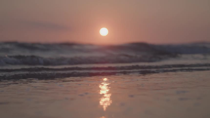 The sun sets low on the horizon casting a shimmering reflection on the wet sand waves roll in and break on the shore their foamy white caps contrasting with the warm glow of the setting sun