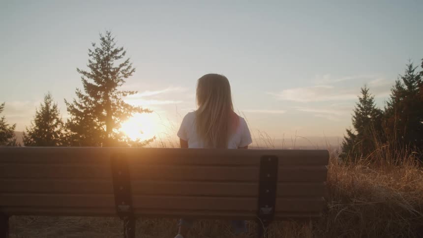 A woman with long hair sits on a bench her back to the camera looking out at a hazy sunset the scene is framed by trees and a golden sunbeam shines through the trees and onto the bench