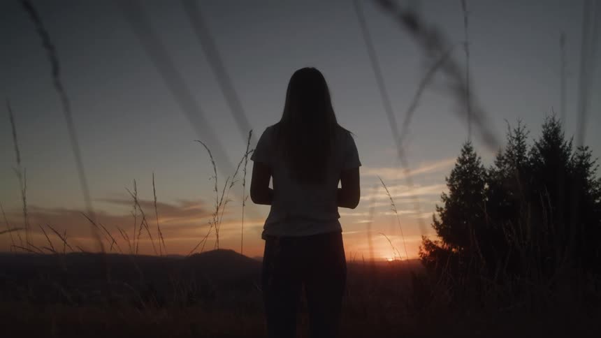 The silhouette of a womanstanding outdoors is illuminated by the setting sun