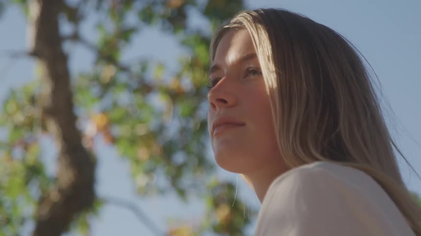 A blonde woman looks upward and to the right with a thoughtful expression on her face her hair is lit by the sun and the background is a blurredout blue sky and green trees