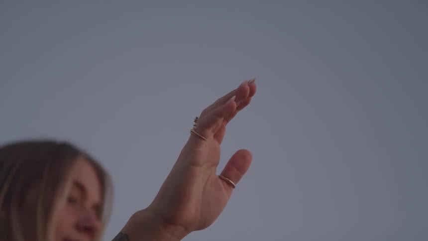 A young woman with a flower tattoo on her forearm and a gold ring on her finger holds her hand up