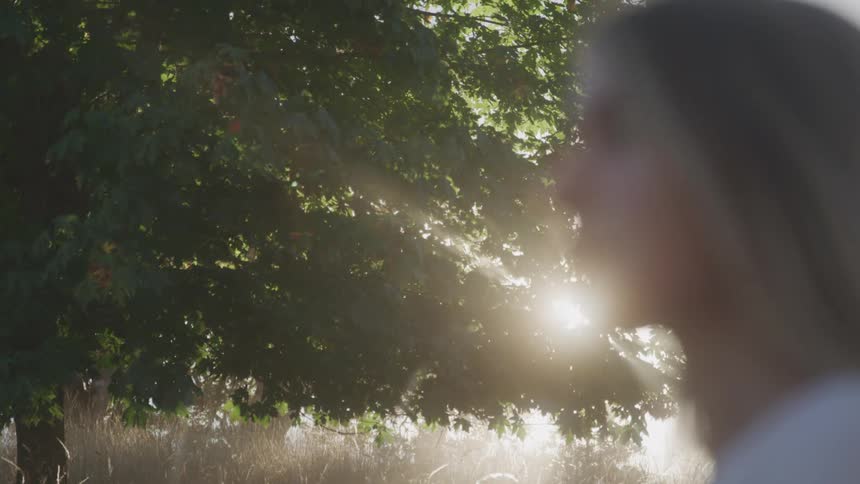 A tree with bright sun rays passing through the leaves in an outdoor environment and in front of it a blonde woman looking forward