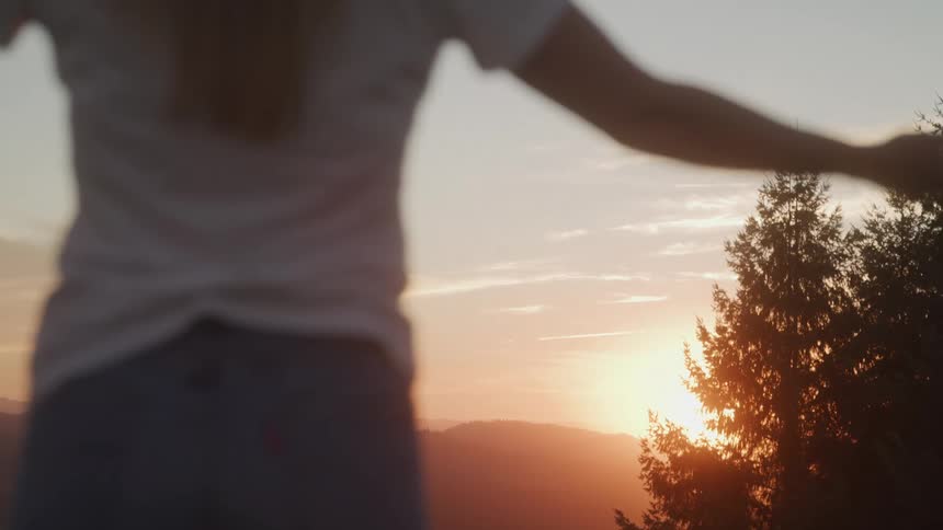 The video shows a person standing on a hill or mountain with their back to the camera looking at the sunset the sky is a beautiful mix of orange yellow and blue hues the sun is visible behind a tall green pine tree with its rays creating a warm g