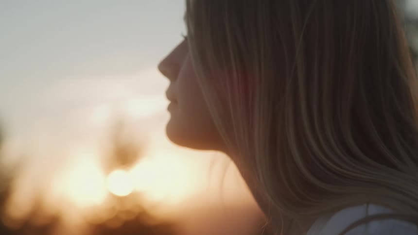 The face of a young blonde woman walking and behind her the sun shining orange in the sky