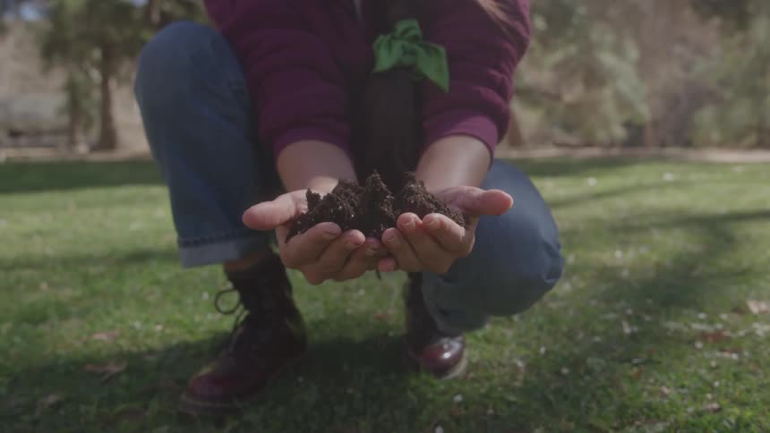 A person wearing brown boots and blue jeans kneels on a grassy lawn holding up a small pile of dark rich soil in their cupped hands