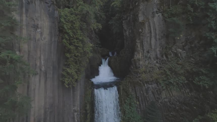A highangle shot shows a waterfall gushing between two towering mossy rock faces the water plummets down into a pool below creating a powerful cascade