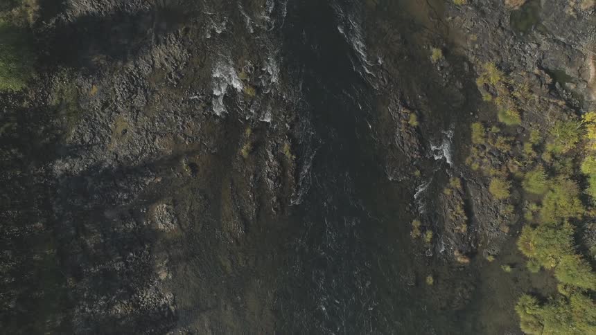 The video shows an overhead drone shot of a river flowing over a rocky bed the water on the left side is choppy and creates small areas of whitewater while the right side of the river is calmer and appears dark the banks are covered in rocks and scatte