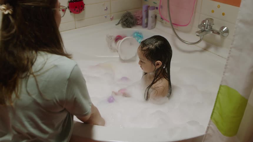 Mother leans into the tub while watching her daughter take a bubble bath