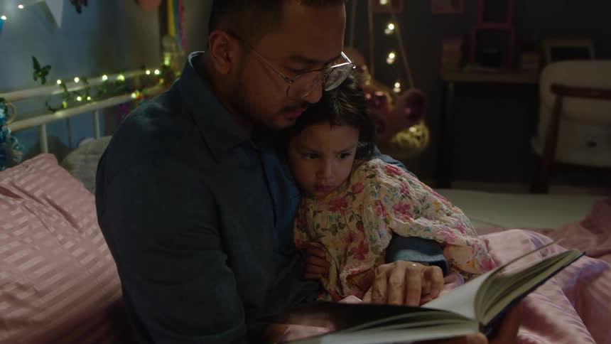 The video shows a man and a young girl sitting together on a bed the man is holding an open book and pointing to something on the page while the girl who is wrapped in a blanket looks on