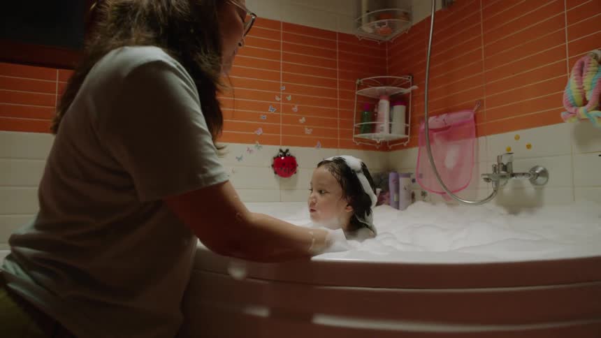 The video shows an adult and a young girl in a brightly lit bathroom taking a bubble bath the adult is playing with the girl creating large playful shapes with the foam the girl is smiling and appears to be enjoying the bath