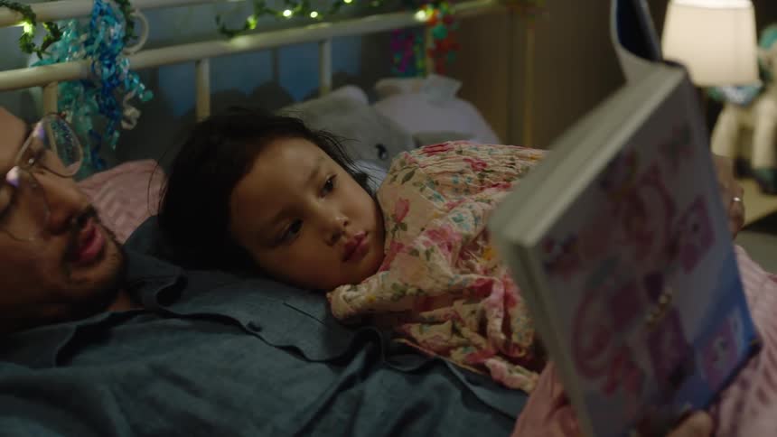 The video shows a child lying on a bed while a person reads to them the camera pans from a wide shot of the bedroom which has fairy lights a unicorn plushie and a teddy bear in a hanging chair to a closeup of the child and the persons face