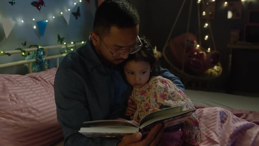 The video shows an intimate moment between a father and his young child in a dimly lit cozy bedroom they are sitting on a bed covered in pink blankets and the father is holding and reading a book to the child who is leaning against him the room is de