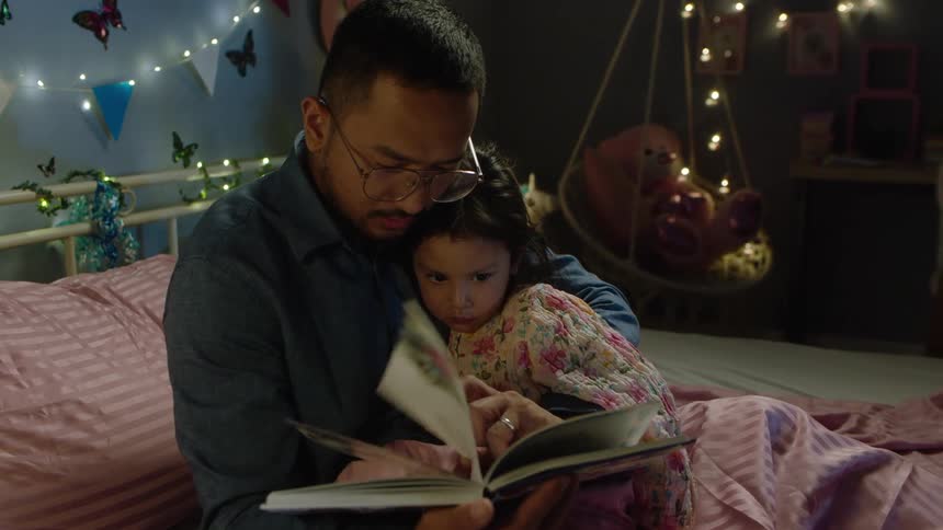 A man and a young child are sitting together on a bed in a bedroom the man is holding a book and reading to the child
