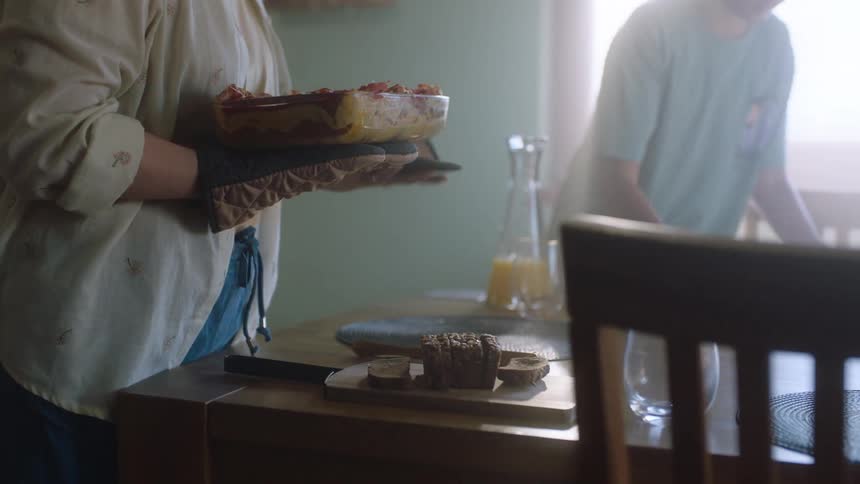 A person wearing oven mitts takes a hot dish out of the oven they then place the dish on a kitchen counter and remove the mitts