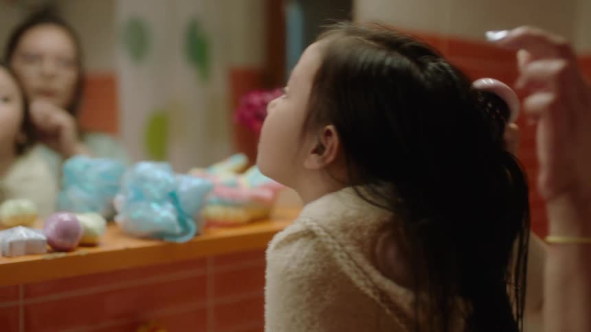 A caring mother brushing her daughters long dark hair in the bathroom as the little girl watches her reflection in the mirror
